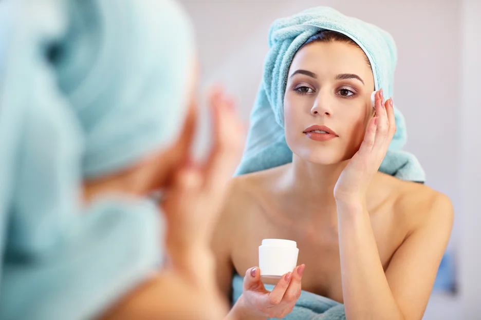 a woman applies moisturizer on her face while looking at the mirror